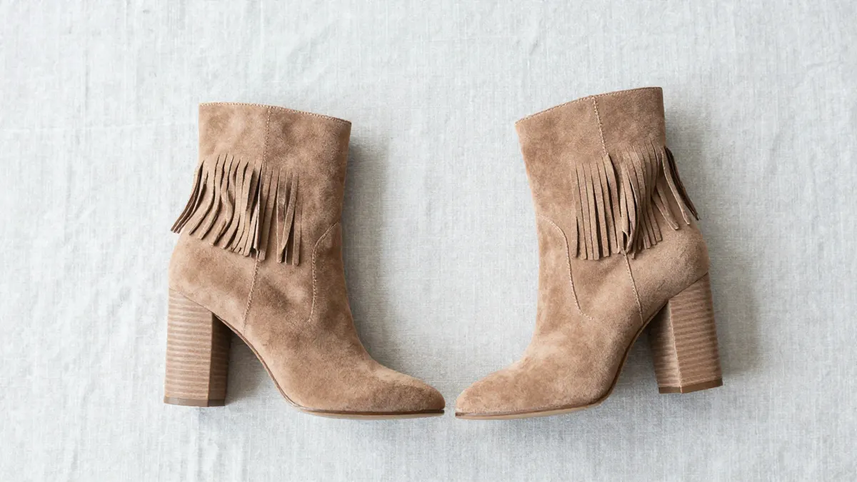 Pair of tan suede peep toe booties with fringe detailing standing on a sunlit wooden floor